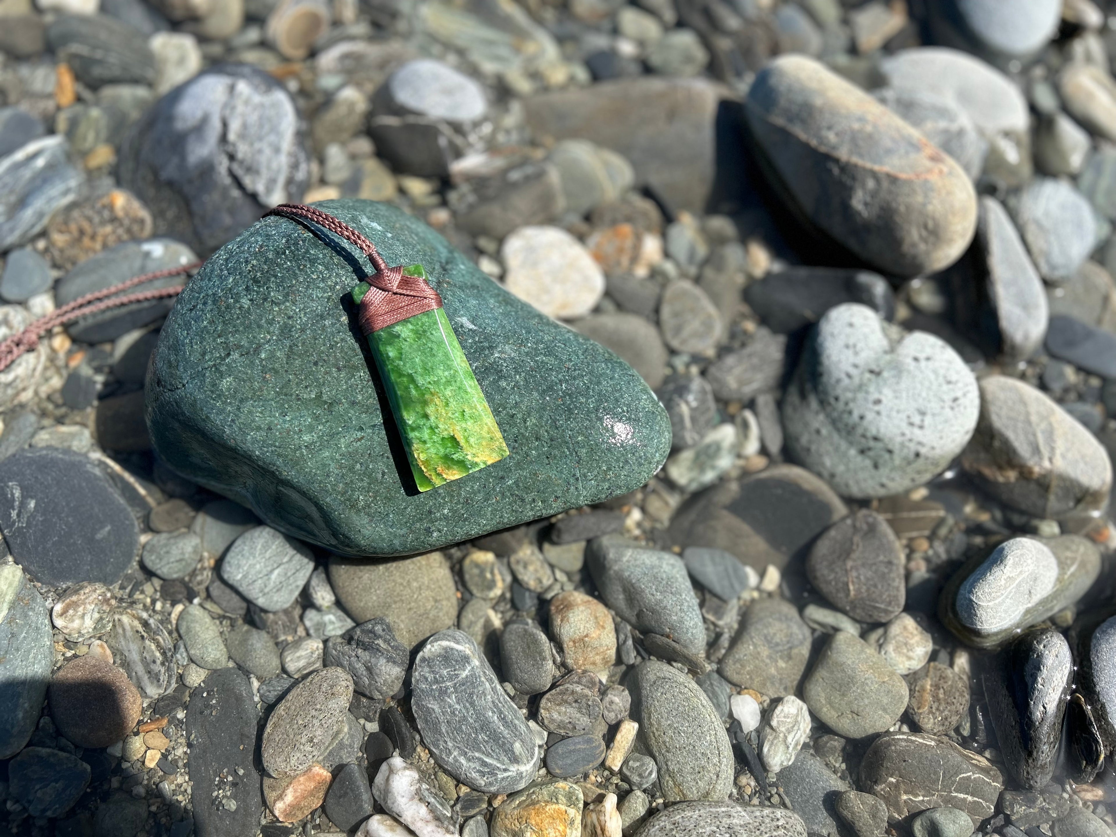 Pounamu (greenstone/jade) toki pendant resting on a rocky riverbed